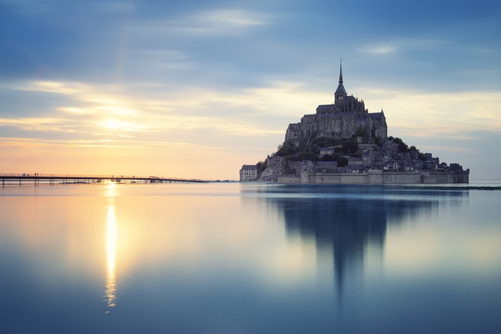 Le Mont-Saint-Michel au crépuscule, se reflétant les eaux calmes qui l'entourent pendant une grande marée, avec le soleil couchant créant des teintes dorées sur l'horizon.